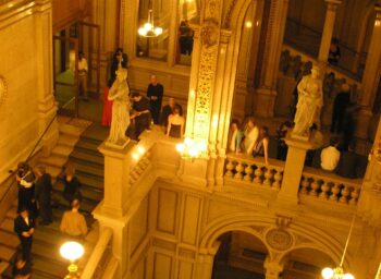 Elegant people in evening dress walk and gather on ornate staircases in a large, warmly lit hall with classical architecture, decorative statues and intricate details on the walls.