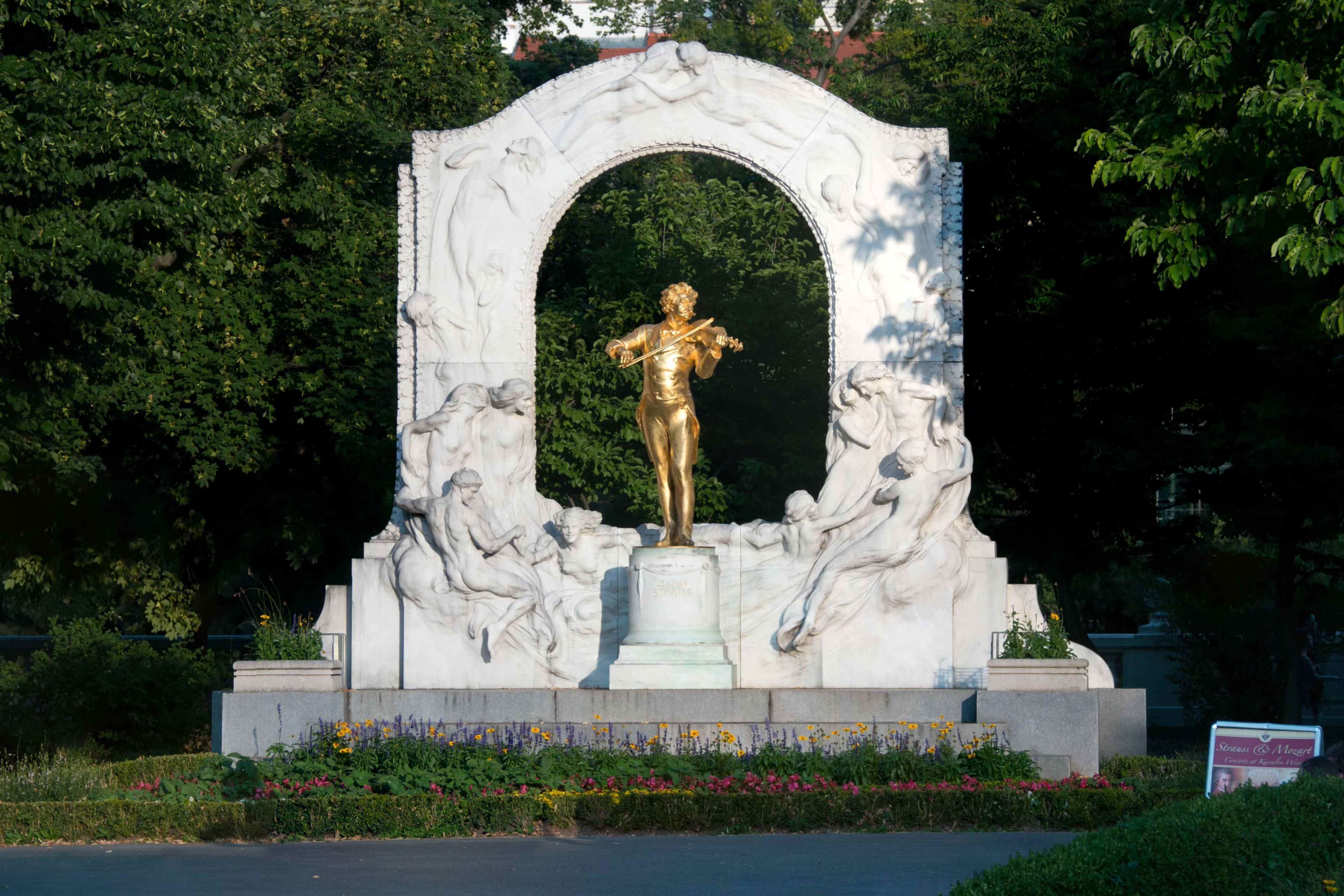 Eine goldene Statue des Geigers Johann Strauss steht unter einem wei&szlig;en Steinbogen, der mit Skulpturen geschm&uuml;ckt ist, umgeben von Gr&uuml;n und bunten Blumen in einer Parklandschaft.