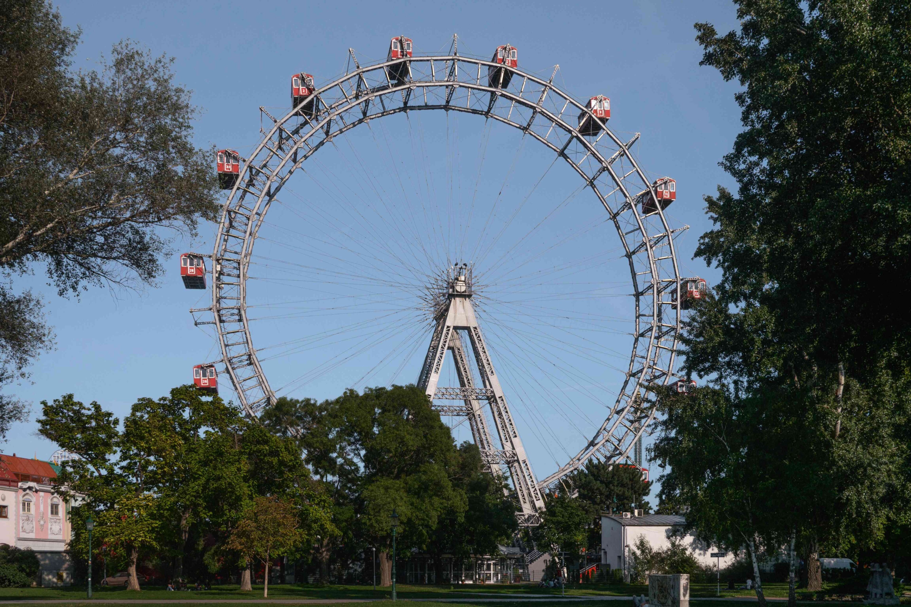 Una grande ruota panoramica con cabine rosse si erge tra alberi e verde sotto un cielo azzurro e limpido.