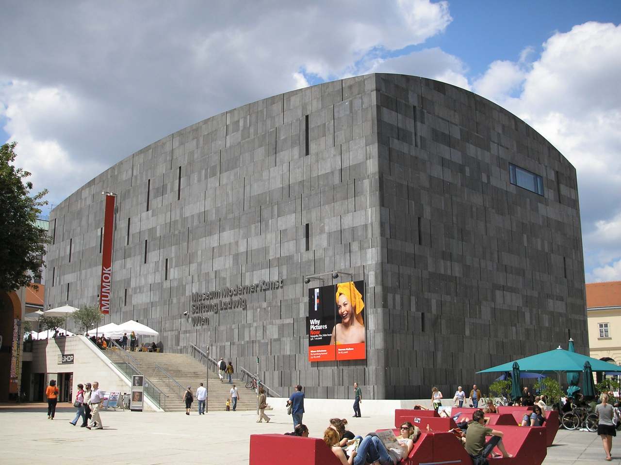 Ein gro&szlig;es, modernes graues Steingeb&auml;ude mit minimalen Fenstern, das als Museum f&uuml;r zeitgen&ouml;ssische Kunst Mumok bezeichnet wird. Menschen sitzen auf roten B&auml;nken auf einem sonnigen Platz mit Regenschirmen und einer Werbetafel davor.