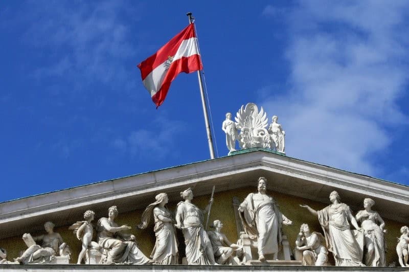 Die österreichische Flagge weht auf einem klassizistischen Gebäude mit detaillierten weißen Statuen und Schnitzereien vor einem strahlend blauen Himmel mit vereinzelten Wolken.