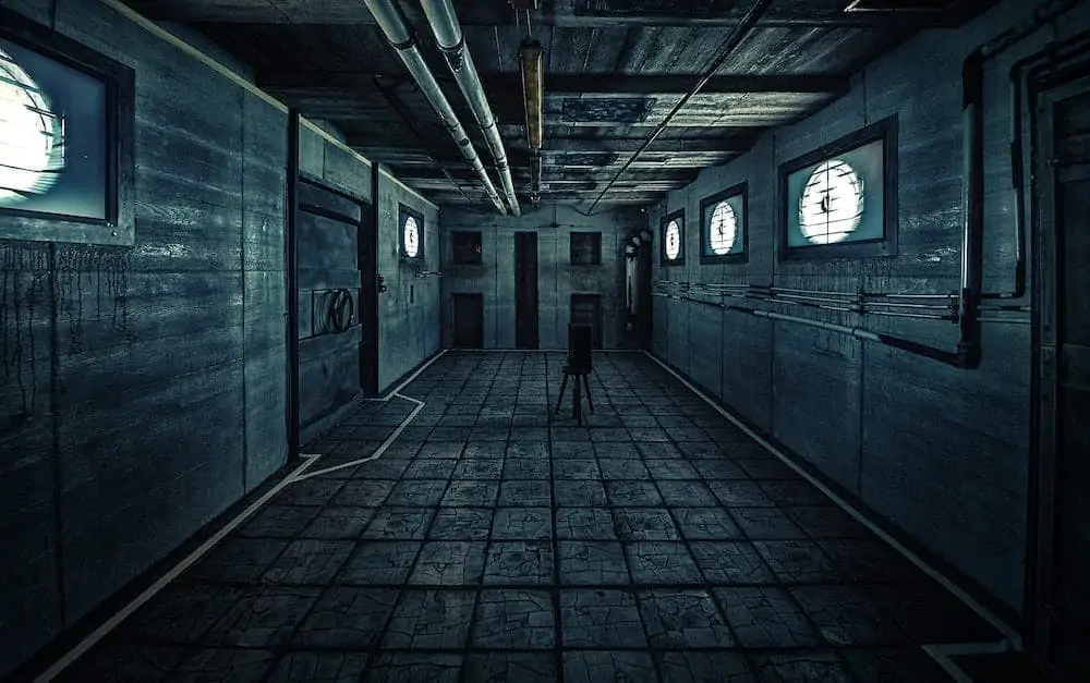 Тускло освещенный, пустой промышленный коридор с бетонными стенами и полом. Круглые лампы на стене отбрасывают холодный свет. Вдоль потолка тянутся трубы, а в центре стоит одинокий стул. Комната выглядит жуткой и пустынной.