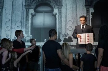 La figura di cera di un uomo che tiene in mano un libro aperto si trova su un palco davanti all'immagine di un edificio storico. Un gruppo di persone, tra cui bambini e adulti, si trova in basso, osserva la scena e la indica.