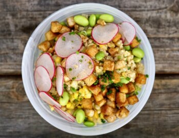Eine Sch&uuml;ssel R&uuml;hrtofu, belegt mit Radieschenscheiben, Edamame und gehacktem Schnittlauch auf einer Holzoberfl&auml;che. Das Gericht besteht aus einer Mischung aus Mais, Reis und gew&uuml;rfelten Tofust&uuml;cken.