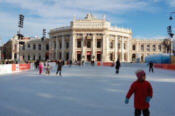 Des personnes profitent du patinage sur une grande patinoire devant un bâtiment historique élaboré avec des colonnes et des statues. Le ciel est clair et bleu et certaines personnes portent des vêtements d'hiver comme des manteaux et des bonnets.