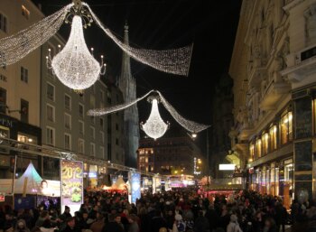 Un affollato mercatino di Natale all'aperto di notte, con luci festose e grandi lampadari. Una folla di persone si muove tra le bancarelle decorate in una strada affollata. Gli edifici sono illuminati e creano un'atmosfera calda e accogliente.