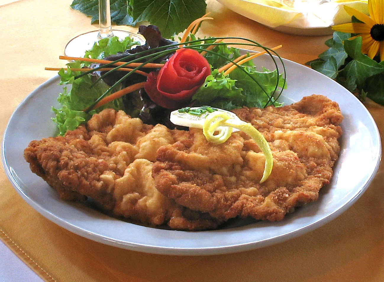 Ein Teller mit paniertem und gebratenem Schnitzel, garniert mit einer Zitronenscheibe und Kräutern, serviert mit gemischtem Grün, Gurke und einer in Form einer Rose geschnittenen Tomate.