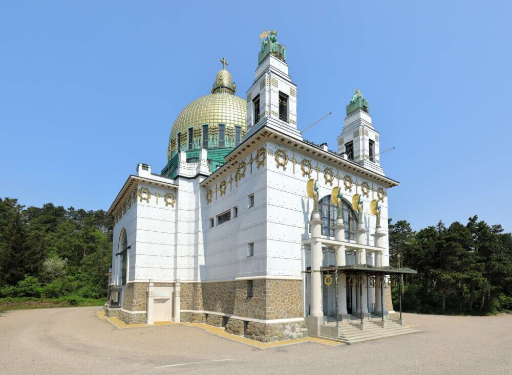 Une église blanche et or avec un grand dôme vert et deux hautes tours, entourée d'arbres sous un ciel bleu clair. L'édifice présente des décorations ornementales et un soubassement en pierre.