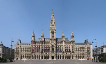 Un magnifique bâtiment néo-gothique avec une tour d'horloge centrale, plusieurs tours et des fenêtres cintrées ornées de drapeaux, qui se dresse sur une vaste place vide sous un ciel bleu limpide.
