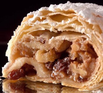 Eine Nahaufnahme eines aufgeschnittenen Apfelstrudels mit einem goldgelben, mit Puderzucker bestäubten Blätterteig und einer Füllung aus Äpfeln, Rosinen und Nüssen.