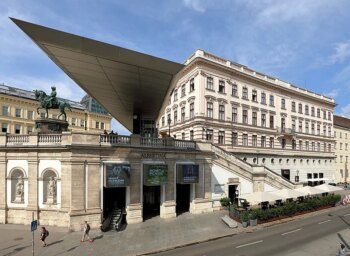 El museo Albertina de Viena (Austria) presenta una gran fachada neoclásica con una gran marquesina moderna de cristal, una estatua ecuestre, escaleras exteriores y una terraza con gente cenando en un café.