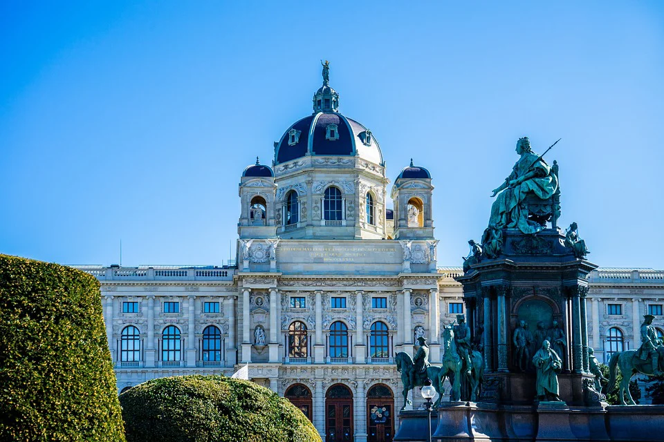 Ein prächtiges historisches Gebäude mit einer großen Kuppel und einer kunstvollen Architektur steht hinter einer Statue mit mehreren Figuren unter einem klaren blauen Himmel mit grünen Sträuchern im Vordergrund.
