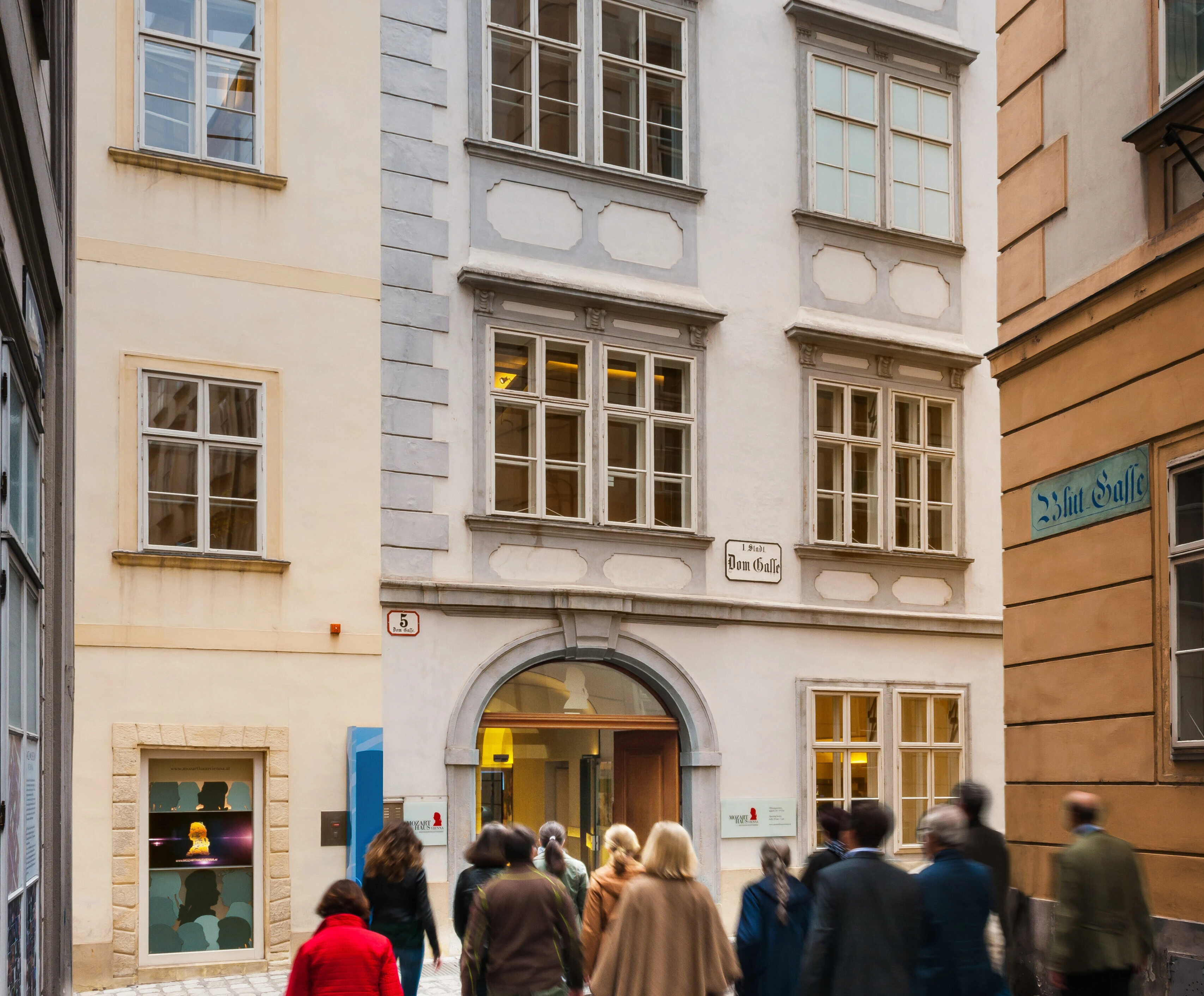 一群人走在一条狭窄的街道上,街道两旁是带有华丽窗户的多层建筑。其中一栋建筑的入口处挂着一块牌子。现在是白天,街上熙熙攘攘。