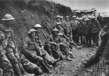 Un groupe de soldats casqués de la Première Guerre mondiale est assis et se tient debout dans une tranchée boueuse, l'air fatigué et solennel. Les murs de la tranchée sont en argile et le ciel au-dessus est couvert. Certains soldats tiennent des fusils et de l'équipement à la main.