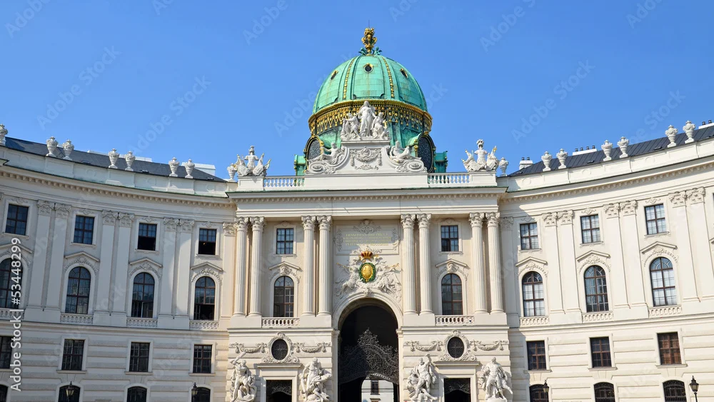 Die reich verzierte Fassade der Hofburg in Wien, Österreich, mit einem zentralen Torbogen, Statuen und einer großen grünen Kuppel mit goldenen Details vor einem klaren blauen Himmel.