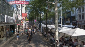 Busy shopping street with lots of people walking and eating under white parasols in street caf&eacute;s. Several store signs, including a large "BORTOLOTTI", are visible. Trees line the pedestrian zone and provide shade.