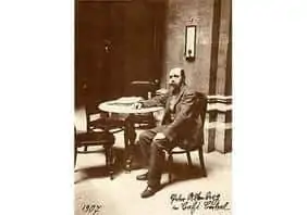 Sepiafarbenes Foto eines b&auml;rtigen Mannes im Anzug, der an einem runden Tisch in einem sp&auml;rlich eingerichteten Raum sitzt, mit einem gerahmten Bild an der Wand hinter ihm und einem handgeschriebenen Text am unteren Rand.