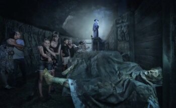 Eine Gruppe von Menschen sieht verängstigt aus, als sie sich in einem schwach beleuchteten, unheimlichen Flur zusammendrängt. Im Hintergrund steht eine Statue in zeitgenössischer Kleidung auf einem Sockel. Im Vordergrund ist eine beunruhigende, leblose Gestalt zu sehen, die auf dem Boden liegt.