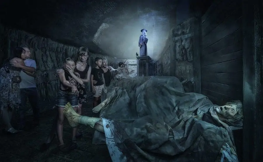 Skupina lidí, kteří se k sobě tisknou v tlumeně osvětlené, strašidelné chodbě, vypadá vyděšeně. V pozadí stojí na podstavci socha v dobovém oblečení. V popředí je vidět znepokojivou postavu bez života ležící na podlaze.