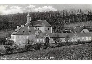 Schwarz-Wei&szlig;-Foto eines gro&szlig;en Anwesens mit mehreren miteinander verbundenen Geb&auml;uden, einem zentralen Bogentor und einer kleinen Kirche mit Kirchturm inmitten eines bewaldeten H&uuml;gels. Am unteren Rand ist deutscher Text zu lesen.