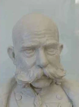 Wykonane z białej gliny lub gipsu popiersie łysego mężczyzny z charakterystycznymi wąsami. Popiersie ma szczegółowe rysy twarzy, w tym głęboko osadzone oczy i poważny wyraz, z płaszczem w stylu wojskowym na piersi.