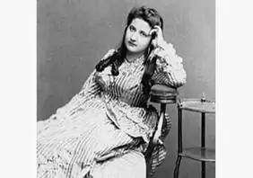 19世紀のストライプの長袖ドレスを着た女性が、小さなテーブルの横に座り、頭を手の上に置いて思案げにカメラを見つめている。.