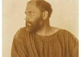 A sepia-toned photo of a bearded man with short hair, wearing a loose, simple garment, looking to his left. The background is plain and light-colored.