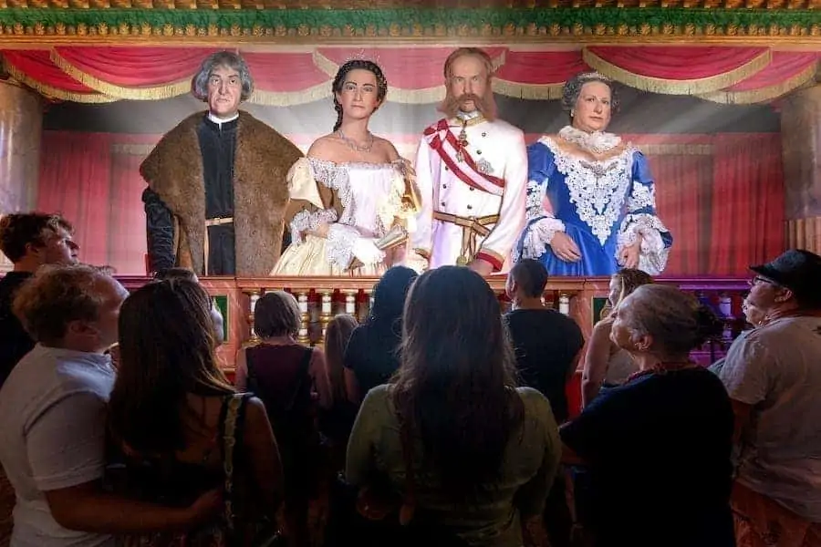 Eine Gruppe von Menschen betrachtet Wachsfiguren in historischen Gewändern auf einer Bühne. Die Figuren stellen Personen aus der Habsburger-Zeit dar und stehen vor einer majestätischen Kulisse mit goldenen Säulen und prächtigen Vorhängen.