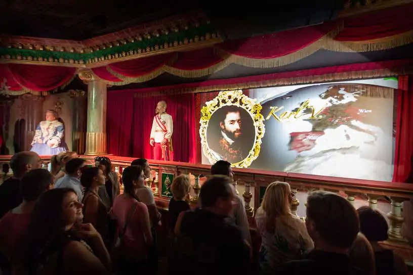 Grupa ludzi ogląda animowaną prezentację historyczną na scenie z ozdobnymi dekoracjami. Na ekranie widać brodatego mężczyznę w ramce obok mapy. Scenę otaczają czerwone zasłony i posągi.