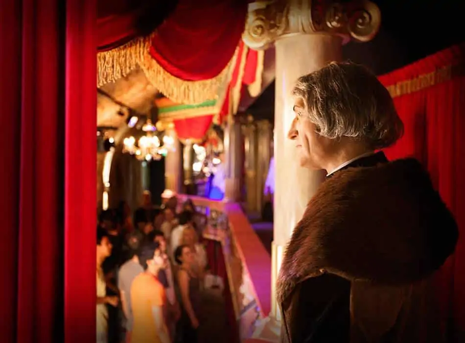 Mężczyzna o siwych włosach, ubrany w ciemną szatę z futrzanym wykończeniem, stoi na balkonie i spogląda na tłum ludzi. Scena jest bogato udekorowana czerwonymi zasłonami, ozdobnymi kolumnami i delikatnym oświetleniem, nadając jej elegancki, historyczny klimat.