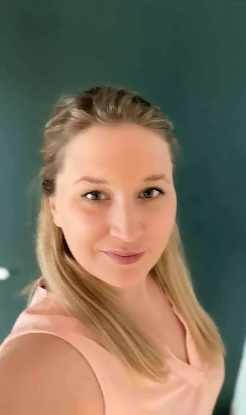 Una persona sonriente con el pelo largo y rubio lleva un top rosa claro y está de pie delante de un fondo verde oscuro.
