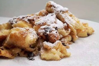 Auf einem weißen Teller liegt ein Haufen goldbrauner, zerrissener Pfannkuchenstücke, die mit Puderzucker bestäubt und mit sichtbaren Schokoladensplittern durchsetzt sind.