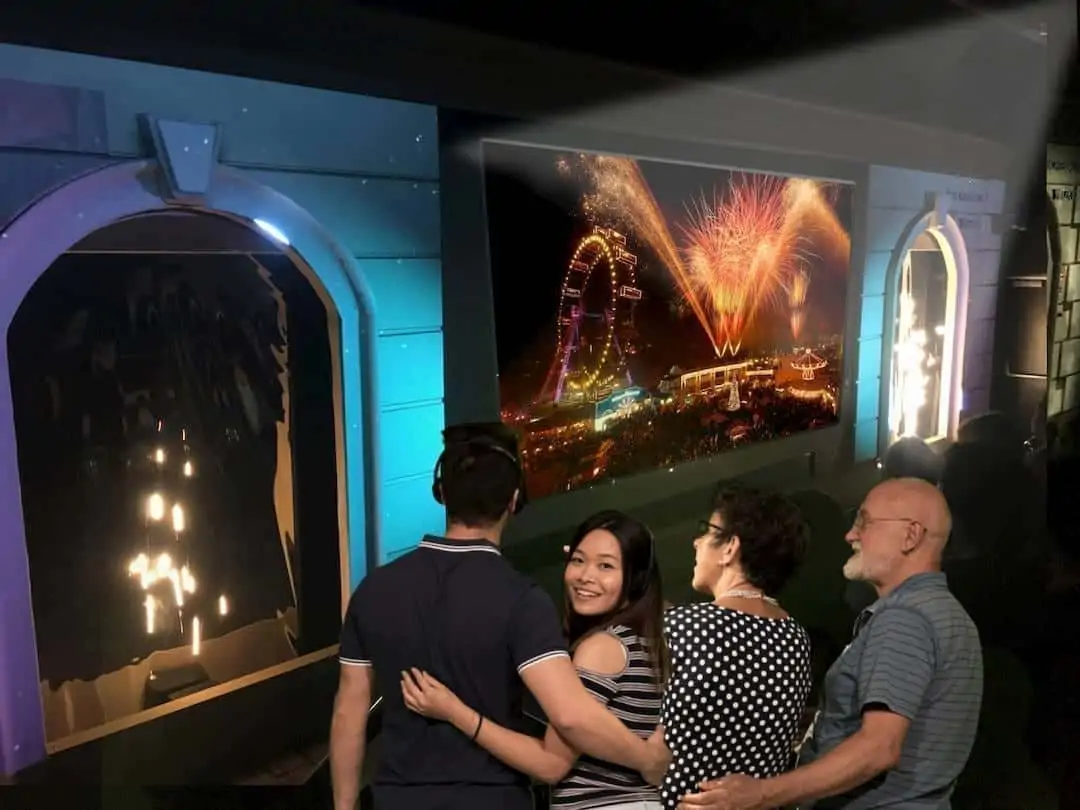 Четыре человека стоят на фоне ярких фейерверков, колеса обозрения и городских огней. Они улыбаются и, кажется, веселятся. Они обнимают друг друга, наблюдая за оживленной сценой.  