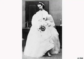 Foto d'epoca in bianco e nero di una persona con un abito bianco elaborato e formale che tiene in mano un bouquet e sta in piedi al chiuso accanto a un tavolo.