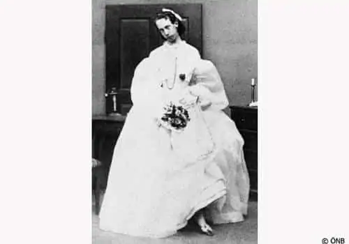 Fekete-fehér fénykép egy személyről, aki egy bő, fehér, viktoriánus stílusú ruhát visel, kezében egy kis csokorral, egy asztal és egy ajtó előtt áll.