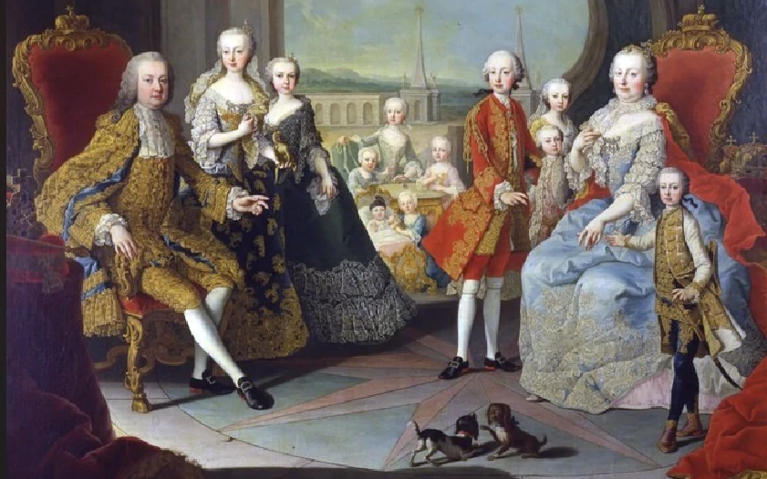 A royal family dressed in elaborate 18th-century clothing poses in a grand room with red chairs, a scenic background, and two small dogs on the floor in the foreground.