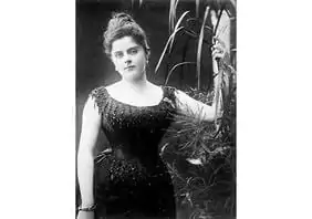 Photographie vintage en noir et blanc d'une femme v&ecirc;tue d'une robe formelle orn&eacute;e de perles, debout &agrave; c&ocirc;t&eacute; de grandes plantes. Elle a les cheveux coiff&eacute;s et regarde directement l'appareil photo avec une expression pos&eacute;e.