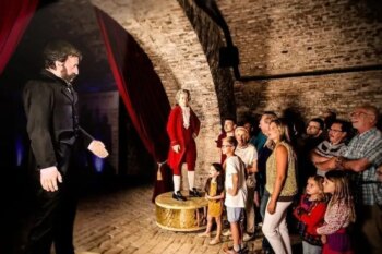 Un gruppo di persone, tra cui bambini e adulti, si trova in una stanza in mattoni a volta poco illuminata. Stanno assistendo a una scena con un uomo in abito nero che indica una statua di un uomo in rosso, forse parte di una rappresentazione storica o teatrale.