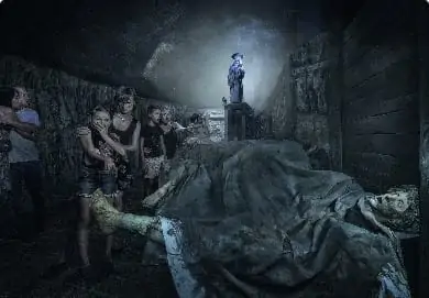 Eine Gruppe von Menschen geht vorsichtig durch einen schwach beleuchteten, unheimlichen Tunnel. Im Hintergrund steht die Statue eines Soldaten. Im Vordergrund ist eine verhüllte Figur auf einer Steinoberfläche zu sehen. Die Atmosphäre ist angespannt und geheimnisvoll.