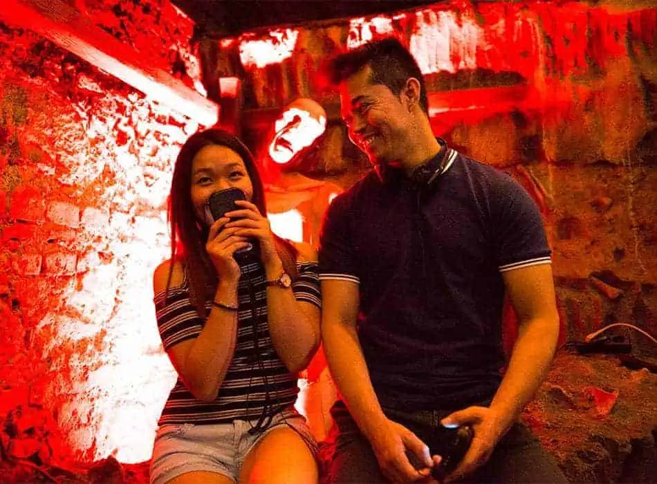 Dva lidé sedí v únikové místnosti s tlumeným červeným osvětlením. Žena vlevo se usmívá a drží mikrofon, muž vpravo se na ni šklebí. V pozadí stojí strašidelná figurína, která umocňuje děsivou atmosféru.