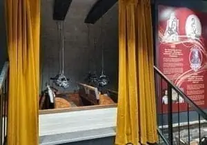 Un groupe de personnes, dont des enfants et des adultes, observe des statues de cire dans une pièce faiblement éclairée aux murs de pierre. Un personnage porte un costume noir, un autre en tenue rouge se tient sur une estrade. Des rideaux rouges encadrent la scène.