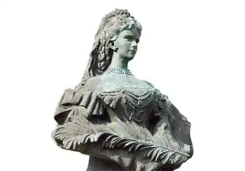 Szczegółowe kamienne popiersie kobiety z wyszukaną fryzurą. Ma na sobie bogato zdobioną suknię z misternymi wzorami i wielorzędowy naszyjnik. Tło jest gładkie, białe.