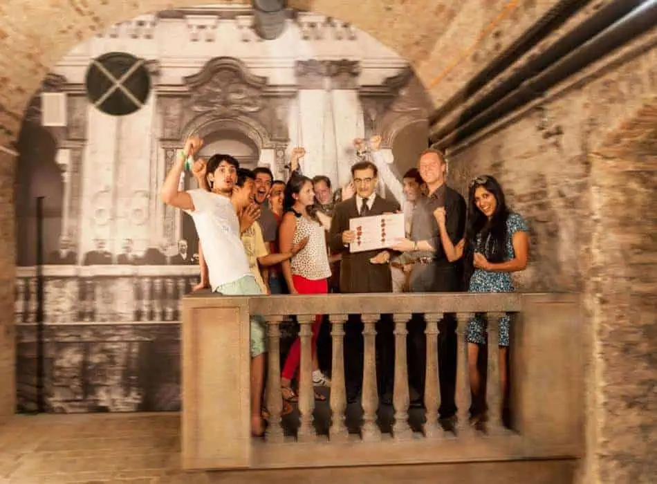 Un gruppo di persone si riunisce eccitato su un balcone, tenendo in mano una scatola davanti a una grande fotografia in bianco e nero di un edificio decorato. La scena sembra essere al chiuso, con pareti di mattoni a vista ed elementi industriali. 