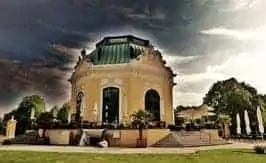 Un edificio hist&oacute;rico redondo, amarillo y con tejado verde se alza bajo un cielo espectacular, rodeado de &aacute;rboles y asientos al aire libre, con escalones que conducen a la entrada.