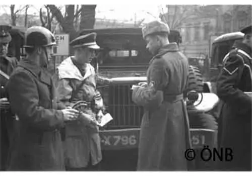 Une photo en noir et blanc montre plusieurs soldats en uniforme debout devant un véhicule militaire, en train de discuter et d'examiner des documents. Des arbres et des bâtiments sont visibles à l'arrière-plan.