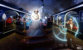 Dans une salle souterraine faiblement &eacute;clair&eacute;e, la statue d'un personnage historique se trouve au centre, entour&eacute;e de personnes assises. &Agrave; droite, une personne appara&icirc;t avec une cam&eacute;ra et des effets de lumi&egrave;re. Le plafond vo&ucirc;t&eacute; donne &agrave; la pi&egrave;ce une atmosph&egrave;re de caverne.  