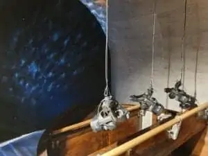 Un orchestre d'hommes en uniforme rouge, dirigé par un chef d'orchestre muni d'une baguette, se produit sur scène. Ils sont assis sur des chaises et jouent des instruments à cordes et des cuivres. Ils tiennent leurs partitions sur des pupitres. L'atmosphère est formelle et concentrée.