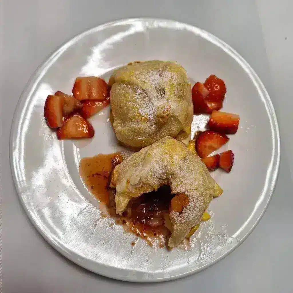 Deux morceaux de pâte partiellement ouverts et saupoudrés de sucre glace, appelés pommes en robe de chambre. Elles sont posées sur une assiette blanche et laissent entrevoir la garniture sucrée. Des tranches de fraises fraîches sont éparpillées par-dessus, créant un contraste coloré.