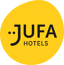 Gelbes, rundes Logo mit der Aufschrift &bdquo;JUFA HOTELS&ldquo; in schwarzem, stilisiertem Text. Die Buchstaben &bdquo;JU&ldquo; sind Teil eines Smiley-Designs.