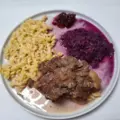 Ein Teller mit Spiralnudeln, geschmortem Rotkohl, einem St&uuml;ck gekochtem Fleisch mit Zwiebeln und einem Klecks Preiselbeerso&szlig;e. Das Gericht wird auf einem hellen Teller serviert, wobei sich etwas So&szlig;e um das Fleisch sammelt.